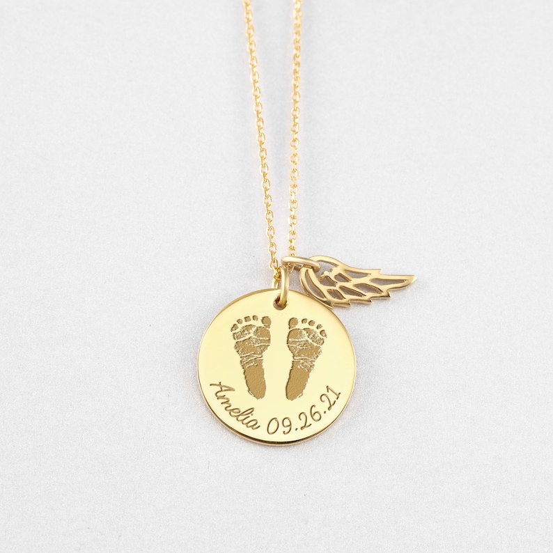 Puede incluir: Un collar dorado con un colgante redondo que presenta dos huellas de beb&eacute; y el nombre "Amelia" con la fecha "09.26.21". Un peque&ntilde;o dije de ala cuelga junto al colgante. El collar est&aacute; sobre un fondo blanco.
