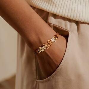 May include: A gold-colored bracelet featuring a floral design. The bracelet is made of a series of connected flower-shaped cutouts. The bracelet is on a brown leather band. The bracelet is worn on a wrist.