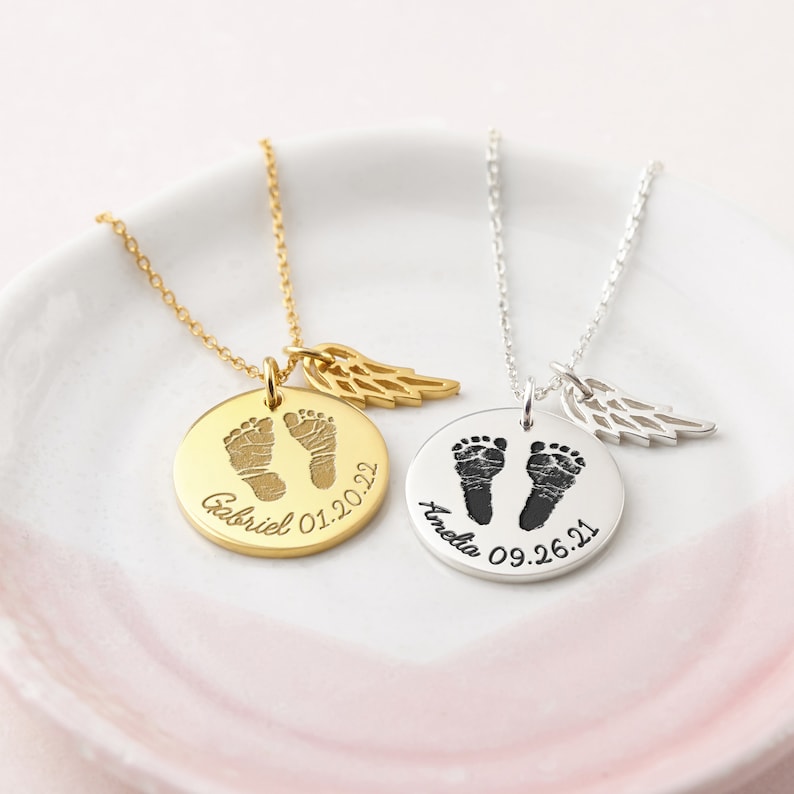 Puede incluir: Dos collares personalizados con colgantes redondos. Uno dorado con huellas de beb&eacute;, el nombre "Gabriel" y la fecha "01.20.22". El otro plateado con "Amelia" y "09.26.21". Cada uno tiene un dije de ala.