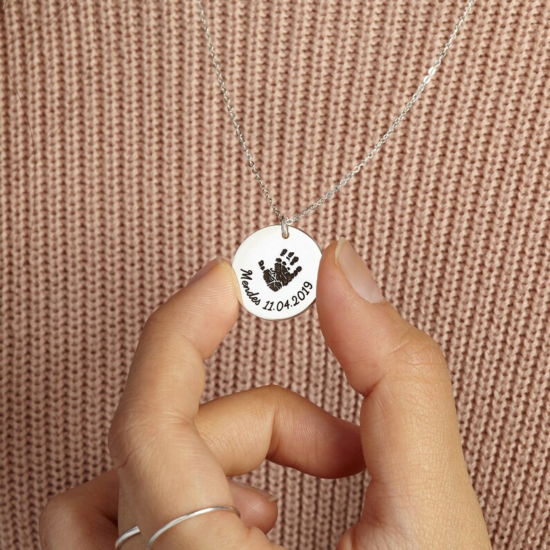 May include: A silver-toned circular pendant necklace featuring an engraved black handprint, the name Mendes, and the date 11.04.2019. The pendant hangs from a delicate silver-toned chain, showcasing a polished metallic finish and detailed engraving.