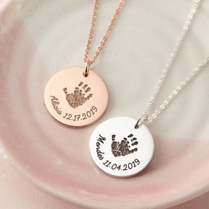 May include: Two circular pendant necklaces, one in rose gold and one in silver, each featuring a dark engraved handprint, a name, and a date. The rose gold pendant is engraved with Alicia 12.17.2019, and the silver pendant is engraved with Mendes 11.04.2019.