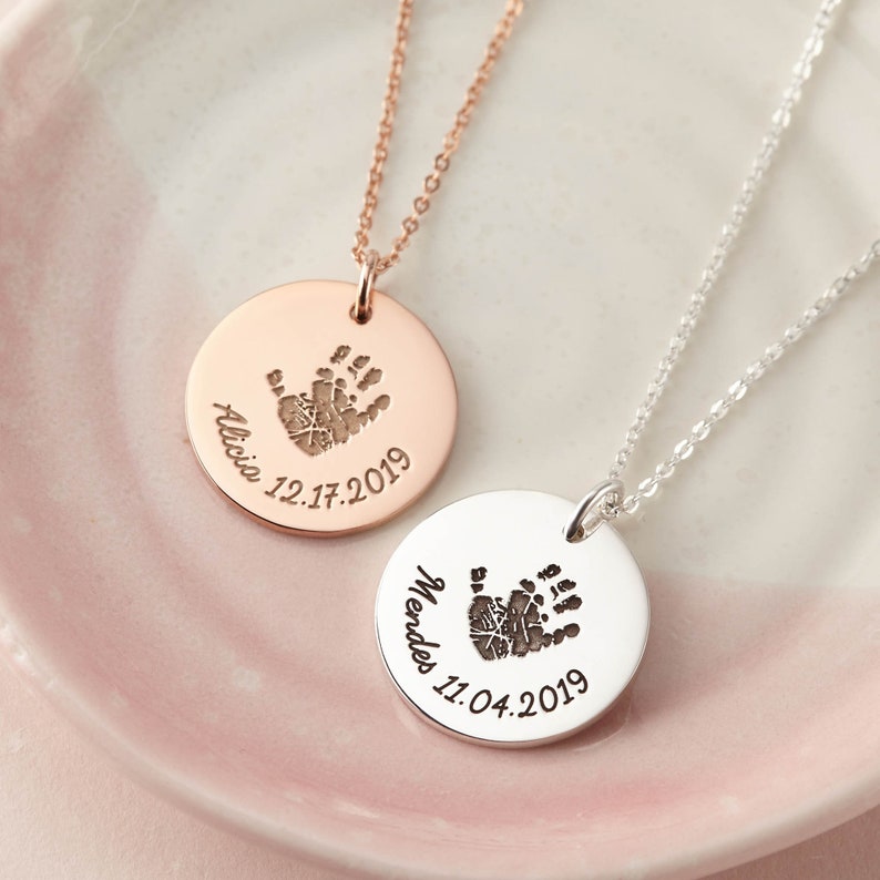 May include: Two circular pendant necklaces, one in rose gold and one in silver, each featuring a dark engraved handprint, a name, and a date. The rose gold pendant is engraved with Alicia 12.17.2019, and the silver pendant is engraved with Mendes 11.04.2019.