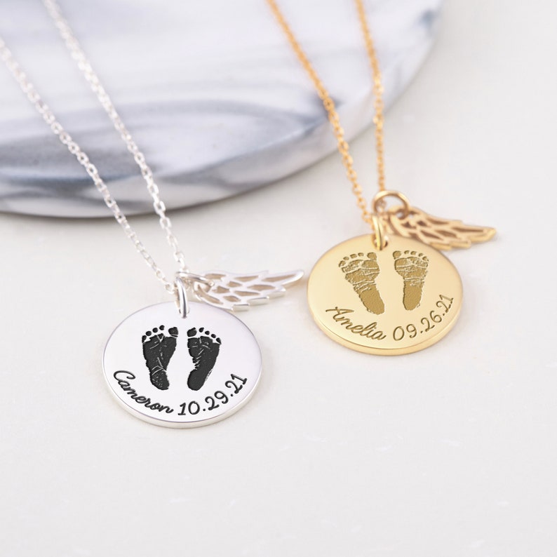 Puede incluir: Dos collares personalizados con huellas de beb&eacute;. Uno es plateado con el nombre "Cameron" y la fecha "10.29.21". El otro es dorado con el nombre "Amelia" y la fecha "09.26.21". Cada collar tiene un peque&ntilde;o dije de ala.