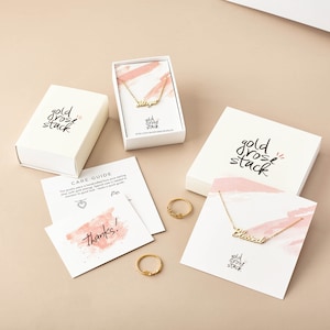 May include: Two gold-tone necklaces with script pendants reading "Blessed" and "Mama" on white cards with pink watercolor accents, two gold-tone rings, two white gift boxes labeled "gold rose stack," and two informational cards with care instructions.