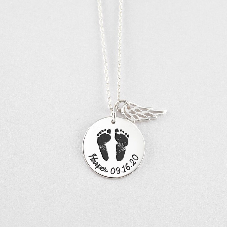 Puede incluir: Un collar de plata con un colgante redondo con huellas de beb&eacute; y el nombre "Harper" con la fecha "09.16.20". Un peque&ntilde;o dije de ala cuelga al lado. El collar est&aacute; sobre un fondo blanco.
