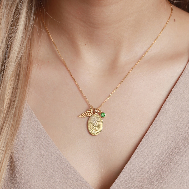 A model wearing a gold fingerprint necklace, complete with an angel wing and green birthstone charm, highlighting its elegant design.