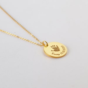 May include: A gold-toned necklace featuring a circular pendant engraved with a small handprint, the name Kacey, and the date 01.18.2020. The pendant is attached to a delicate gold-toned chain, creating a simple and elegant piece of jewelry.