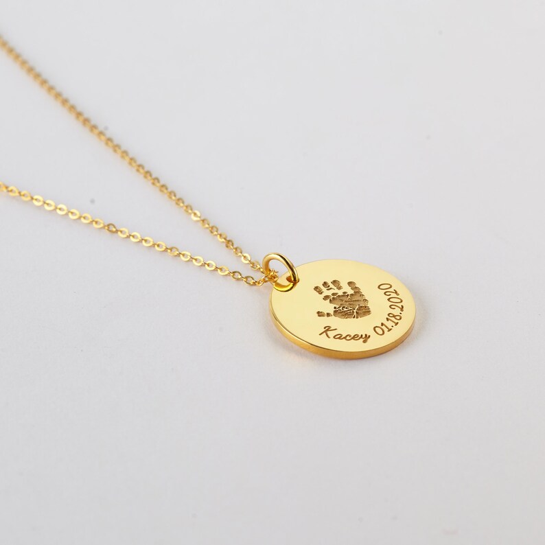 May include: A gold-toned necklace featuring a circular pendant engraved with a small handprint, the name Kacey, and the date 01.18.2020. The pendant is attached to a delicate gold-toned chain, creating a simple and elegant piece of jewelry.