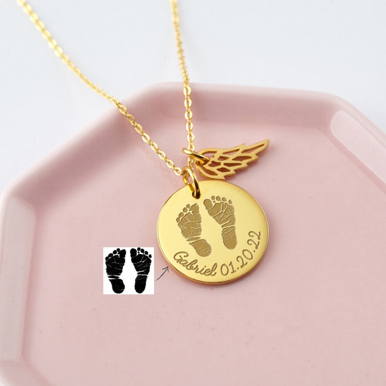 Puede incluir: Un collar dorado con un colgante redondo con huellas de beb&eacute;, el nombre "Gabriel" y la fecha "01.20.22". Un peque&ntilde;o dije de ala cuelga al lado. El collar descansa sobre una superficie rosa.