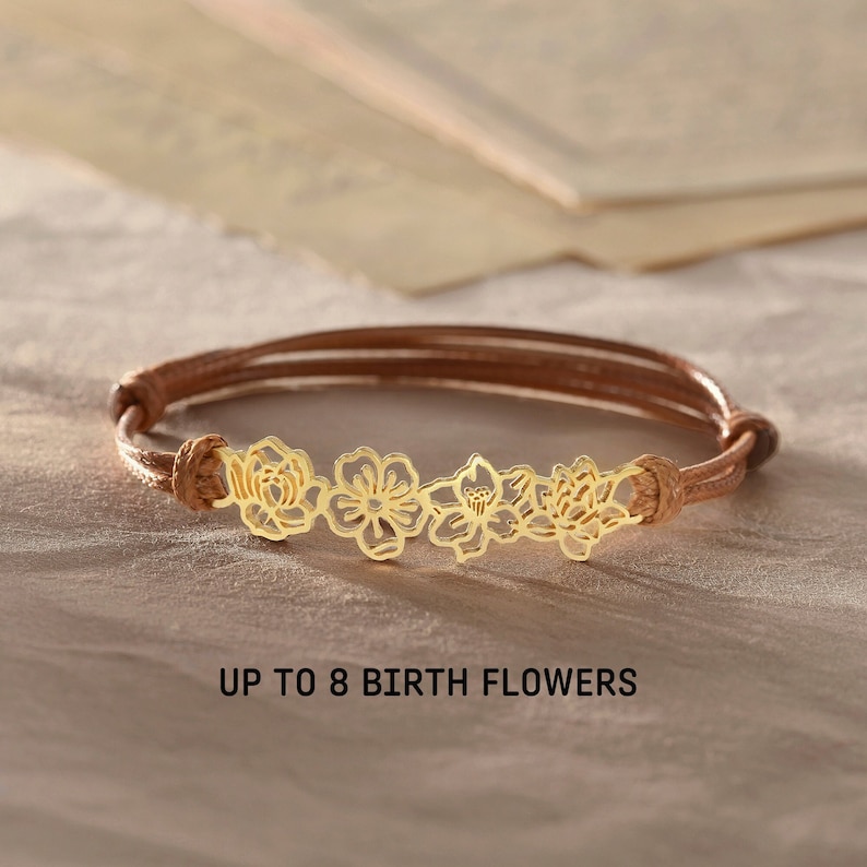May include: A brown leather bracelet with a gold-colored floral design. The bracelet features a series of delicate flower cutouts. The text "UP TO 8 BIRTH FLOWERS" is displayed below the bracelet.