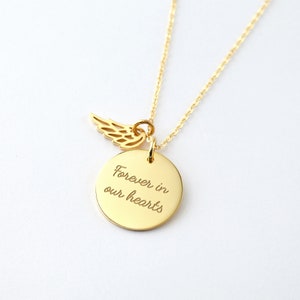 Puede incluir: Un collar dorado con una cadena fina. El colgante presenta un disco redondo grabado con las palabras "Forever in our hearts" y un peque&ntilde;o dije de ala. El collar se muestra sobre un fondo blanco.