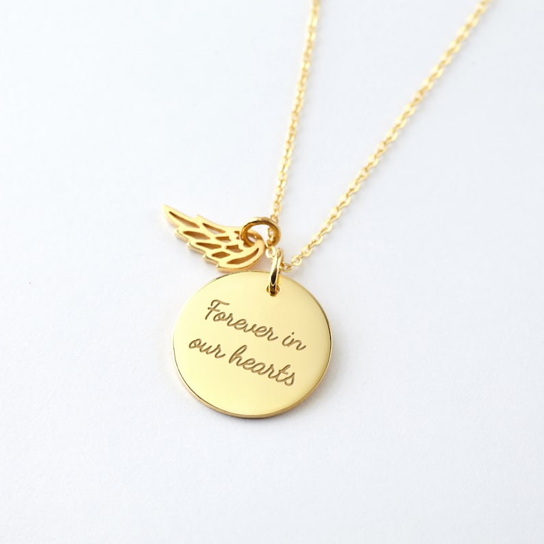 Puede incluir: Un collar dorado con una cadena fina. El colgante presenta un disco redondo grabado con las palabras "Forever in our hearts" y un peque&ntilde;o dije de ala. El collar se muestra sobre un fondo blanco.