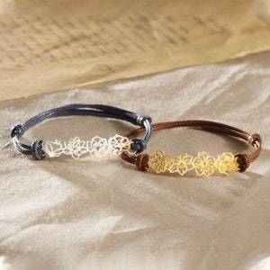 May include: Two bracelets with floral designs. One bracelet has a silver-colored floral design on a dark blue cord. The other bracelet has a gold-colored floral design on a brown cord. Both bracelets are adjustable.