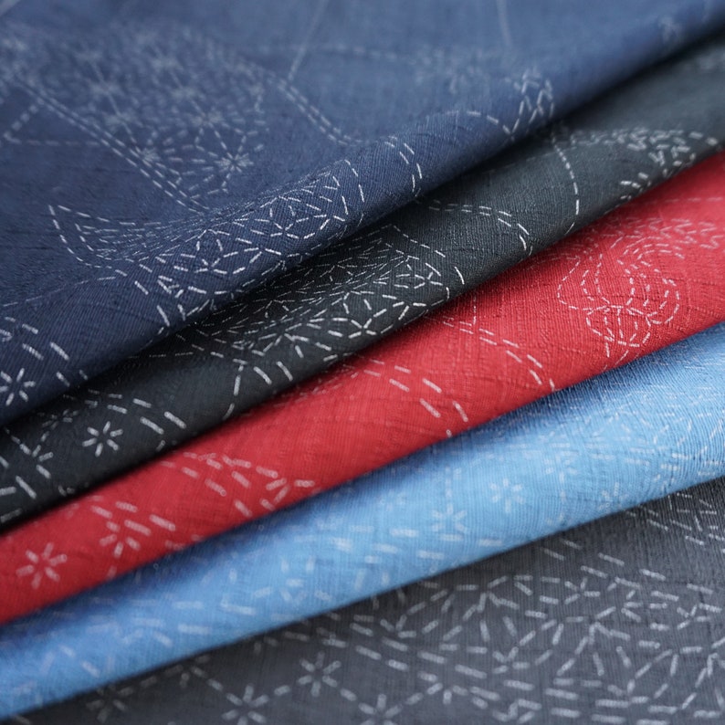 May include: Four fabric squares in different colors, navy blue, black, red, and light blue, are stacked on top of each other. Each square has a white embroidered design of a repeating pattern.