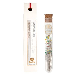 May include: A glass tube filled with colourful patchwork pins. The tube has a cork stopper and a label that reads "Patchwork Pins". The tube is sitting on a white background.