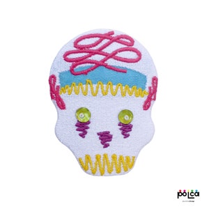 Puede incluir: Parche de calavera bordado en blanco con detalles en rosa, amarillo, azul y verde. La calavera tiene un remolino rosa en la parte superior, líneas amarillas en zigzag alrededor de la parte inferior y ojos de lentejuelas verdes.