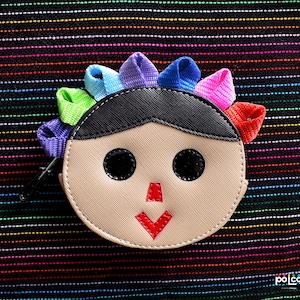 May include: A small, round, beige coin purse with a smiling face. The purse has black eyes, a red nose, and a red mouth. The purse is decorated with a rainbow of colourful fabric strips around the top.