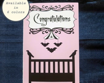 May include: A pink card with black lettering that says "Congratulations". The card features a black silhouette of a crib with a mobile above it. The mobile has a spider web and bats hanging from it.