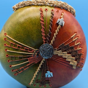 Könnte beinhalten: Handgefertigte Kürbis-Kunst mit Farbverlauf von Grün nach Rot. Verziert mit aufwendigen Nähten, geflochtenen Kordeln und Metallverzierungen, darunter ein zentraler Knopf und Vogelanhänger. Der Rand ist mit einer gewebten Textur versehen.
