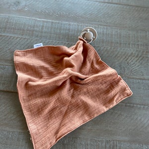 Peut inclure: Une couverture de s&eacute;curit&eacute; carr&eacute;e en mousseline de couleur rouille, dot&eacute;e d'une texture froiss&eacute;e douce et d'une petite &eacute;tiquette en tissu blanc sur le c&ocirc;t&eacute;. Une sucette gris clair est fix&eacute;e &agrave; un coin par une boucle en tissu assortie.