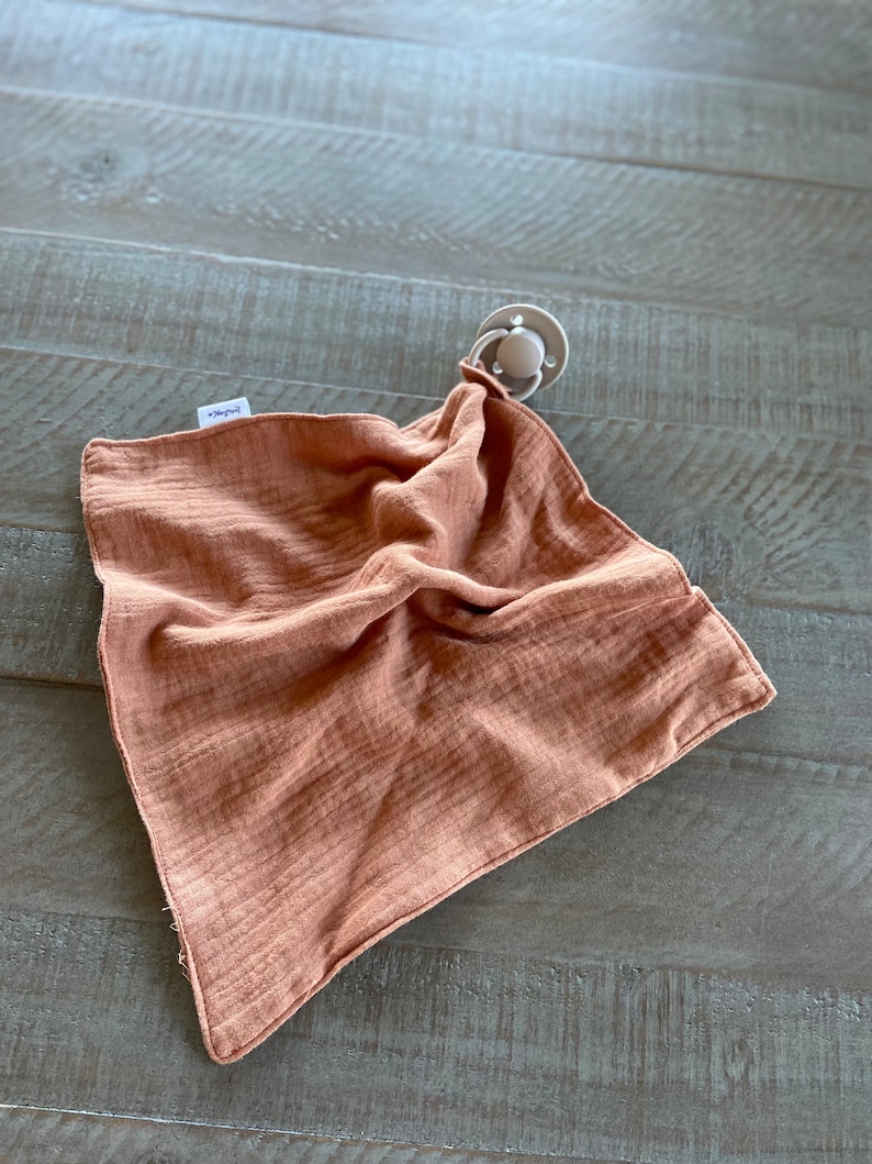 Peut inclure: Une couverture de s&eacute;curit&eacute; carr&eacute;e en mousseline de couleur rouille, dot&eacute;e d'une texture froiss&eacute;e douce et d'une petite &eacute;tiquette en tissu blanc sur le c&ocirc;t&eacute;. Une sucette gris clair est fix&eacute;e &agrave; un coin par une boucle en tissu assortie.