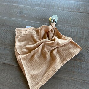 Peut inclure: Une couverture de s&eacute;curit&eacute; en mousseline de couleur beige avec une texture douce et froiss&eacute;e. Le tissu est rassembl&eacute; &agrave; un coin et attach&eacute; solidement &agrave; une sucette vert sauge clair avec une collerette ronde et un embout jaune p&acirc;le.