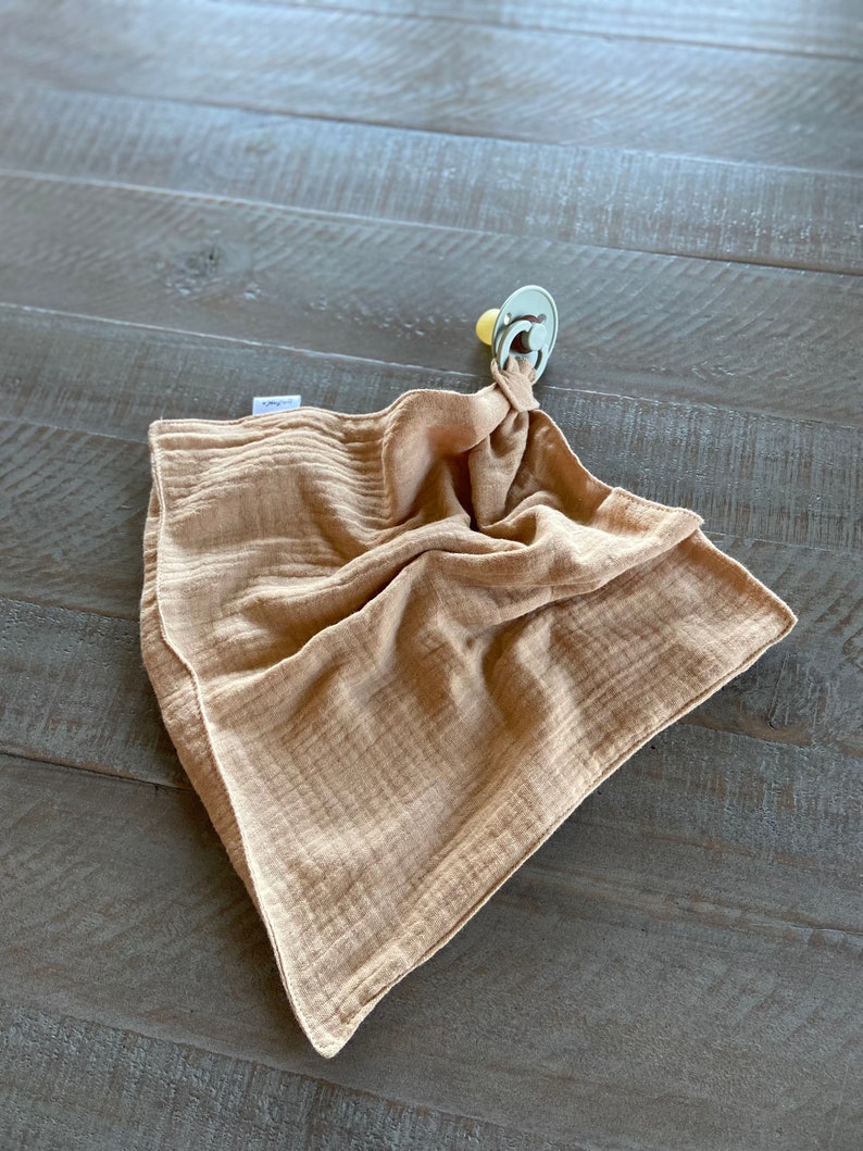 Peut inclure: Une couverture de s&eacute;curit&eacute; en mousseline de couleur beige avec une texture douce et froiss&eacute;e. Le tissu est rassembl&eacute; &agrave; un coin et attach&eacute; solidement &agrave; une sucette vert sauge clair avec une collerette ronde et un embout jaune p&acirc;le.