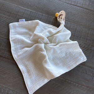 Peut inclure: Une couverture de s&eacute;curit&eacute; carr&eacute;e en mousseline blanc cass&eacute; avec une texture douce et froiss&eacute;e et une petite &eacute;tiquette en tissu sur le c&ocirc;t&eacute;. Une sucette beige avec une t&eacute;tine ronde est fix&eacute;e &agrave; un coin par une boucle en tissu nou&eacute;e.