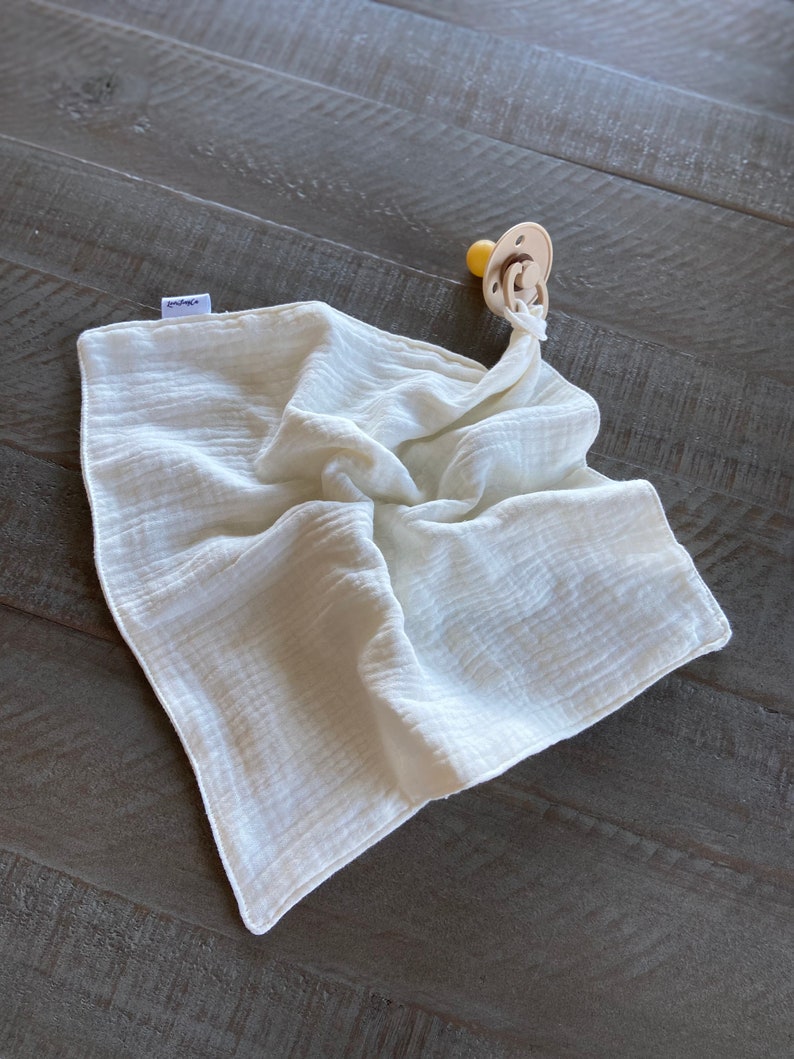 Peut inclure: Une couverture de s&eacute;curit&eacute; carr&eacute;e en mousseline blanc cass&eacute; avec une texture douce et froiss&eacute;e et une petite &eacute;tiquette en tissu sur le c&ocirc;t&eacute;. Une sucette beige avec une t&eacute;tine ronde est fix&eacute;e &agrave; un coin par une boucle en tissu nou&eacute;e.