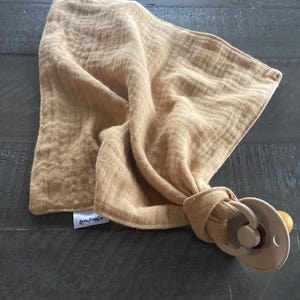Peut inclure: Un carr&eacute; de mousseline jaune moutarde &agrave; la texture froiss&eacute;e, dot&eacute; d'une petite &eacute;tiquette en tissu blanc avec le texte noir "LeeTinyCo". Le tissu est nou&eacute; autour d'une sucette en plastique beige avec une collerette ronde et une t&eacute;tine.