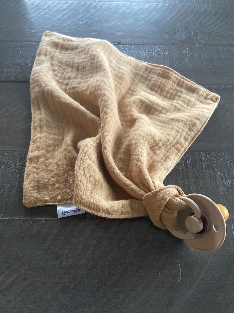 Peut inclure: Un carr&eacute; de mousseline jaune moutarde &agrave; la texture froiss&eacute;e, dot&eacute; d'une petite &eacute;tiquette en tissu blanc avec le texte noir "LeeTinyCo". Le tissu est nou&eacute; autour d'une sucette en plastique beige avec une collerette ronde et une t&eacute;tine.