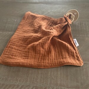 Peut inclure: Une couverture de s&eacute;curit&eacute; en mousseline couleur rouille avec une texture douce et froiss&eacute;e, dot&eacute;e d'une sucette beige attach&eacute;e &agrave; un coin. Une petite &eacute;tiquette en tissu blanc avec du texte noir est cousue sur le c&ocirc;t&eacute; du tissu carr&eacute; aux bords cousus.