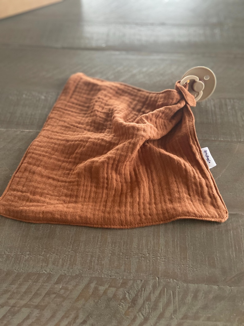 Peut inclure: Une couverture de s&eacute;curit&eacute; en mousseline couleur rouille avec une texture douce et froiss&eacute;e, dot&eacute;e d'une sucette beige attach&eacute;e &agrave; un coin. Une petite &eacute;tiquette en tissu blanc avec du texte noir est cousue sur le c&ocirc;t&eacute; du tissu carr&eacute; aux bords cousus.