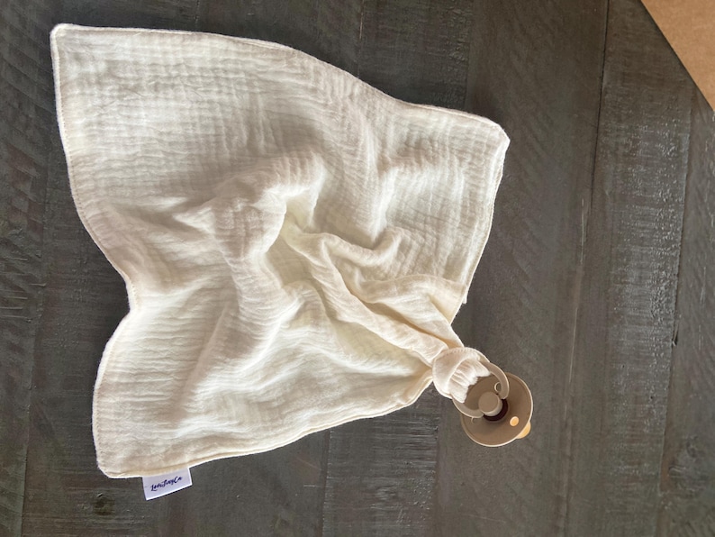 Peut inclure: Un carr&eacute; de mousseline couleur cr&egrave;me &agrave; la texture douce et froiss&eacute;e, dot&eacute; d'une petite &eacute;tiquette en tissu blanc dans le coin. Le tissu est fronc&eacute; &agrave; une extr&eacute;mit&eacute; et attach&eacute; &agrave; une sucette en plastique beige avec un anneau circulaire.