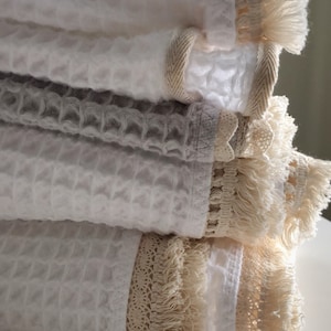 Puede incluir: Una pila de toallas plegadas de tejido de gofre en blanco y gris. Cada toalla está adornada con un borde decorativo y flecos en un color neutro. Las toallas están apiladas, mostrando su textura y diseño.