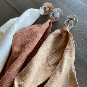 Peut inclure: Trois attaches-sucettes avec des tissus en mousseline assortis en blanc, terracotta et beige. Chaque attache pr&eacute;sente un bouclier en plastique rond avec un anneau central, reli&eacute; &agrave; un carr&eacute; de tissu l&eacute;ger, froiss&eacute; et doux par une boucle nou&eacute;e.