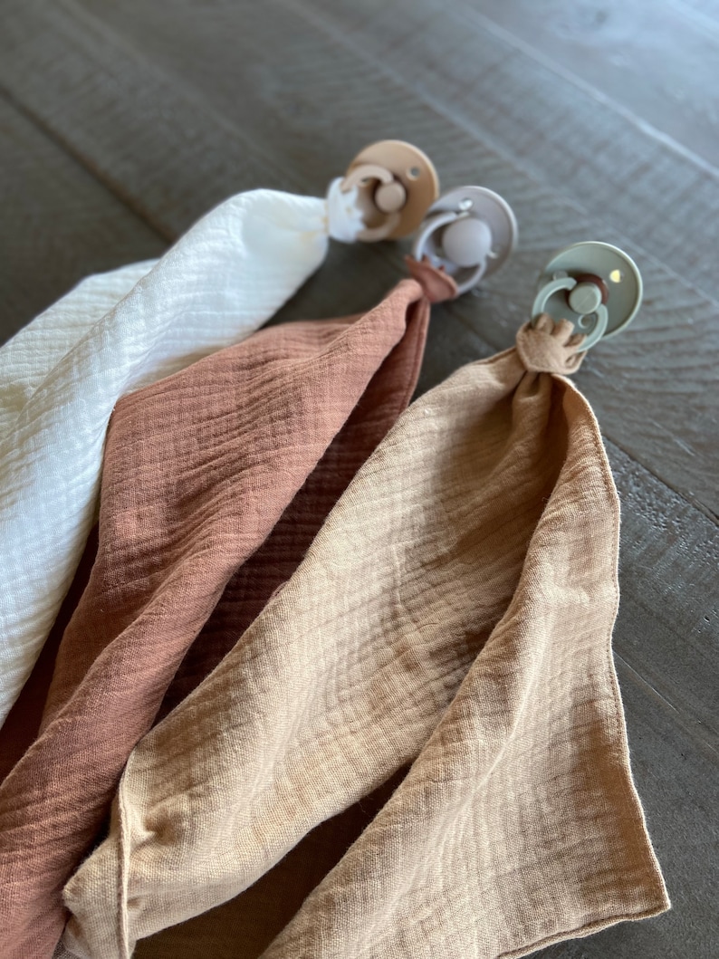 Peut inclure: Trois attaches-sucettes avec des tissus en mousseline assortis en blanc, terracotta et beige. Chaque attache pr&eacute;sente un bouclier en plastique rond avec un anneau central, reli&eacute; &agrave; un carr&eacute; de tissu l&eacute;ger, froiss&eacute; et doux par une boucle nou&eacute;e.