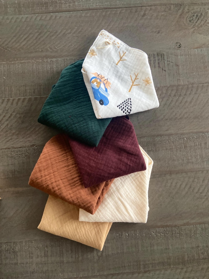 Peut inclure: Un ensemble de six langes en mousseline pli&eacute;s dans des tons vert fonc&eacute;, bordeaux, orange br&ucirc;l&eacute;, jaune moutarde, cr&egrave;me, et un lange blanc orn&eacute; d'un motif de voiture bleue avec des arbres et des formes g&eacute;om&eacute;triques.