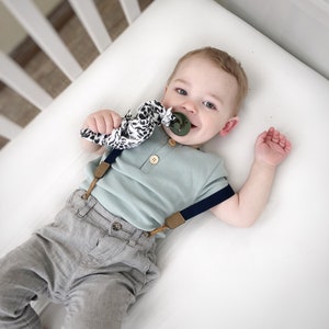 Peut inclure: Une chemise &agrave; manches courtes vert sauge clair avec des boutons en bois, un pantalon gris et des bretelles bleu marine avec des d&eacute;tails en cuir beige. Une sucette vert olive fonc&eacute; est attach&eacute;e &agrave; une attache-sucette en tissu &agrave; motif animal.