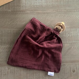Peut inclure: Une couverture de s&eacute;curit&eacute; en mousseline bordeaux fonc&eacute; avec une texture douce et froiss&eacute;e et une petite &eacute;tiquette en tissu blanc dans le coin inf&eacute;rieur. Une sucette beige est fix&eacute;e au coin sup&eacute;rieur du tissu carr&eacute; par une boucle.