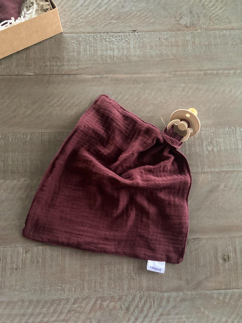 Peut inclure: Une couverture de s&eacute;curit&eacute; en mousseline bordeaux fonc&eacute; avec une texture douce et froiss&eacute;e et une petite &eacute;tiquette en tissu blanc dans le coin inf&eacute;rieur. Une sucette beige est fix&eacute;e au coin sup&eacute;rieur du tissu carr&eacute; par une boucle.