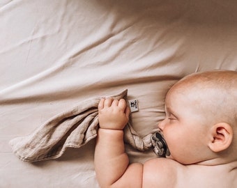 Può includere: Una copertina di sicurezza in mussola beige con una piccola etichetta in tessuto bianco con un logo nero, tenuta da un neonato che dorme con un ciuccio grigio in bocca. La copertina ha una consistenza morbida e stropicciata color marrone chiaro.