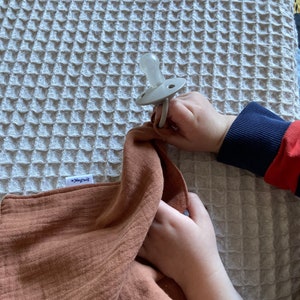 Peut inclure: Une sucette en silicone beige clair avec une collerette ronde et un anneau, attach&eacute;e &agrave; un lange en mousseline couleur rouille dot&eacute; d'une petite &eacute;tiquette blanche portant l'inscription "Little Dutch" en &eacute;criture cursive, sur une couverture gaufr&eacute;e.