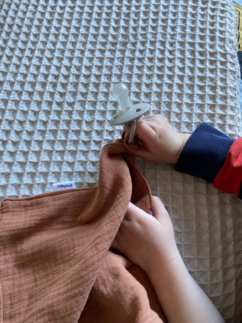 Peut inclure: Une sucette en silicone beige clair avec une collerette ronde et un anneau, attach&eacute;e &agrave; un lange en mousseline couleur rouille dot&eacute; d'une petite &eacute;tiquette blanche portant l'inscription "Little Dutch" en &eacute;criture cursive, sur une couverture gaufr&eacute;e.
