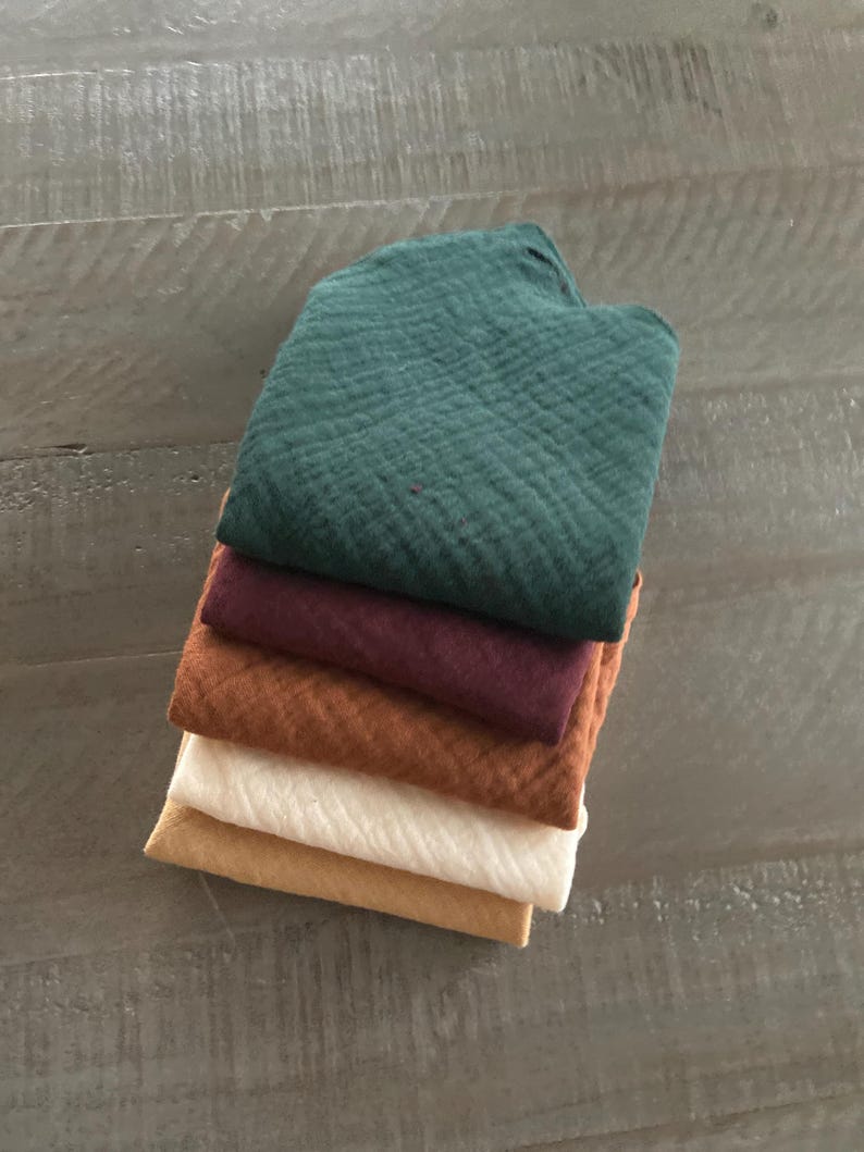 Peut inclure: Une pile de cinq tissus carr&eacute;s pli&eacute;s pr&eacute;sentant un tissage en mousseline textur&eacute; et froiss&eacute;. Les couleurs, de haut en bas, sont le vert for&ecirc;t fonc&eacute;, le bordeaux profond, l'orange br&ucirc;l&eacute;, le blanc cass&eacute; et le jaune moutarde dor&eacute;.