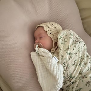 Peut inclure: Un nourrisson endormi portant un bonnet en crochet couleur cr&egrave;me et une sucette, envelopp&eacute; dans une couverture d'emmaillotage en mousseline blanche &agrave; motif floral vert et marron. Le nourrisson repose sur un oreiller textur&eacute; rose p&acirc;le.