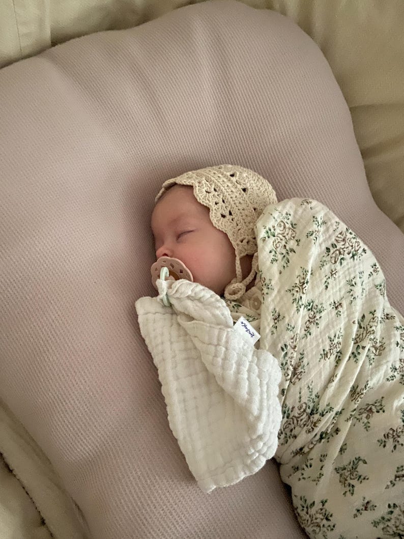 Peut inclure: Un nourrisson endormi portant un bonnet en crochet couleur cr&egrave;me et une sucette, envelopp&eacute; dans une couverture d'emmaillotage en mousseline blanche &agrave; motif floral vert et marron. Le nourrisson repose sur un oreiller textur&eacute; rose p&acirc;le.