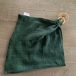 Peut inclure: Une couverture de s&eacute;curit&eacute; carr&eacute;e en mousseline vert for&ecirc;t fonc&eacute; avec une texture froiss&eacute;e. Le tissu est nou&eacute; &agrave; un coin pour fixer une sucette ronde de couleur beige avec un anneau circulaire et une petite &eacute;tiquette blanche de marque.