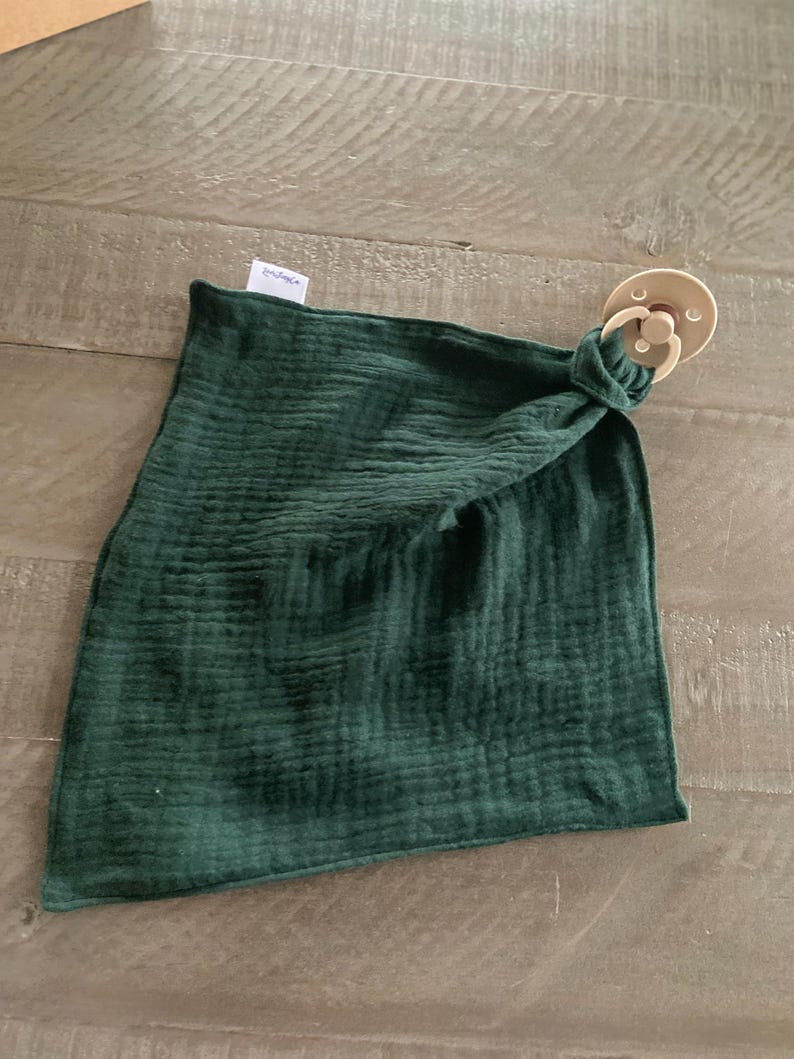 Peut inclure: Une couverture de s&eacute;curit&eacute; carr&eacute;e en mousseline vert for&ecirc;t fonc&eacute; avec une texture froiss&eacute;e. Le tissu est nou&eacute; &agrave; un coin pour fixer une sucette ronde de couleur beige avec un anneau circulaire et une petite &eacute;tiquette blanche de marque.