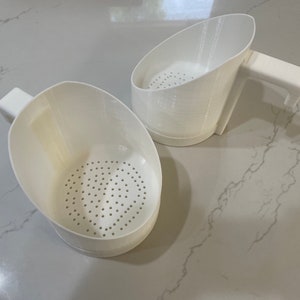 May include: Two white plastic kitchen strainers with handles. The strainers have a round, perforated bottom and a curved, angled top.